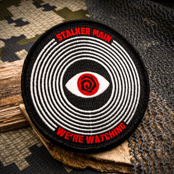 Circular embroidered tactical patch with a hypnotic white spiral design, a red eye in the center, and red embroidered text reading Stalker Main and We're Watchi
