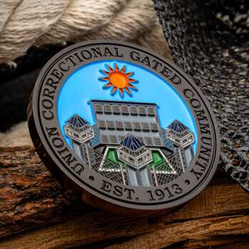 Union Correctional Gated Community challenge coin depicting a facility under a bright orange sun against a blue sky, set on a rustic wooden log with rope.