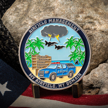 Airfield Management challenge coin featuring a tropical beach design with palm trees, a storm cloud over two fighter jets, and an AMOPS vehicle.