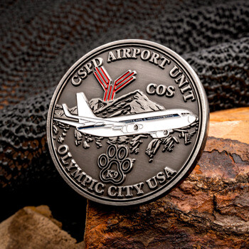 CSPD Airport Unit challenge coin with an airplane, mountain range, red runways, and a K9 paw print, resting on bark.