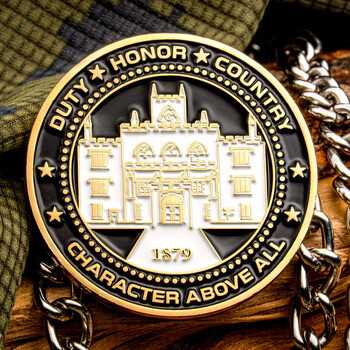 Gold and black enamel military academy challenge coin displaying Duty Honor Country and Character Above All alongside a chain and camouflage fabric.
