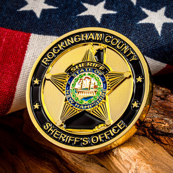 Rockingham County Sheriff's Office challenge coin with a gold sheriff star and State of New Hampshire seal, displayed on wood before an American flag.