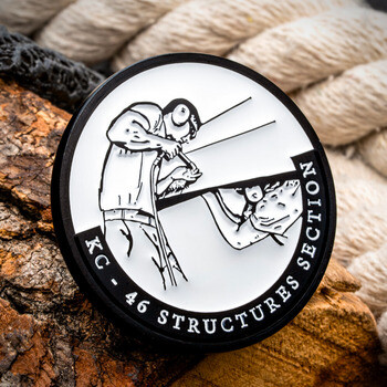 Black and white challenge coin depicting technicians working on aircraft parts with text reading KC - 46 Structures Section.