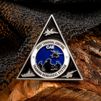Triangular challenge coin for CAE Joint Terminal Control Training Rehearsal System displayed on a wood log with camouflage netting.