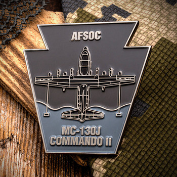 Keystone-shaped challenge coin featuring AFSOC MC-130J Commando II aircraft illustration on a camouflage and wood background.