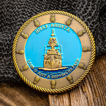 Round brass challenge coin featuring HMAS Arunta ship on a blue background surrounded by pineapple icons and a rope edge.