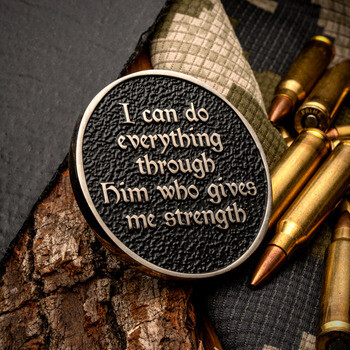 Challenge coin inscribed with scripture next to brass rifle cartridges on camo fabric and tree bark.