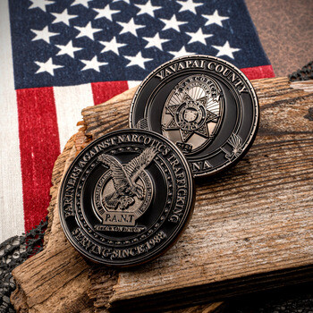 Custom black and silver challenge coins for Yavapai County and Partners Against Narcotics Trafficking, displayed on wood with an American flag background.